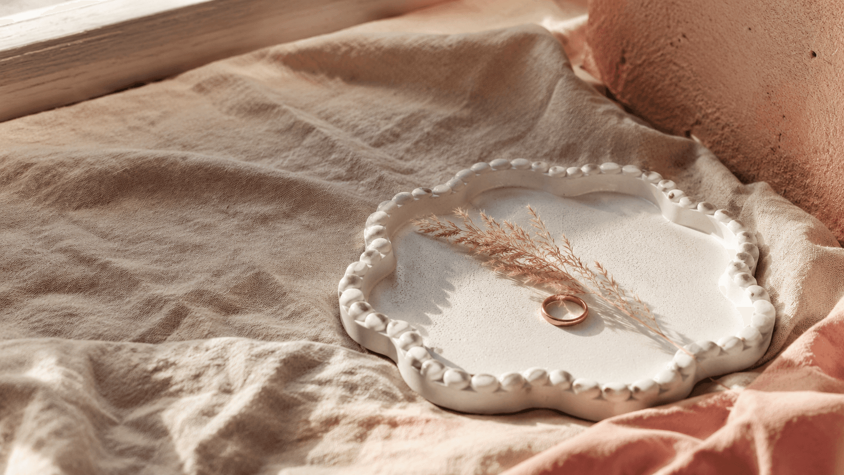 A hand-poured tray with a dried grass sprig and a gold ring, resting on linen